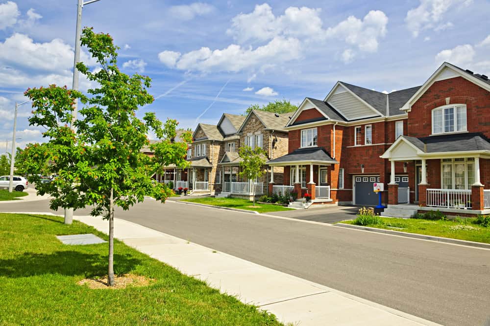 Suburban street with row of brick houses, small front yards, and a sidewalk. Managed by attentive property managers, a young tree thrives near the curb under a partly cloudy sky.