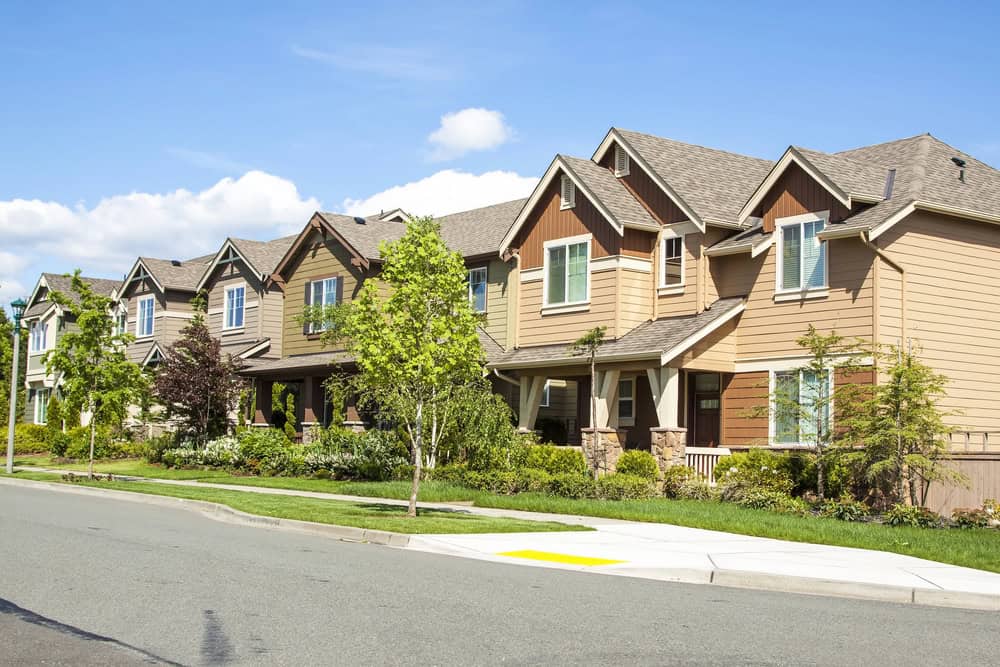 A row of suburban houses with gabled roofs lines the sidewalk, enhanced by lush trees on a sunny day, creating a picturesque community often overseen by diligent property managers.