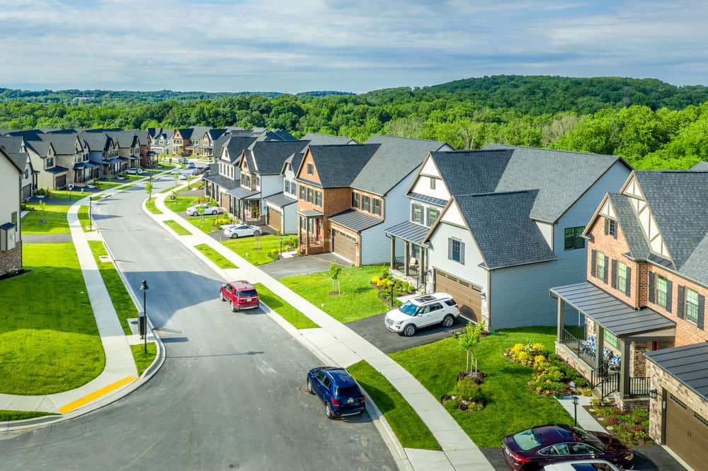 Suburban neighborhood with modern two-story homes, cars parked in driveways, and well-maintained lawns managed by diligent property managers. Aerial view reveals a winding street and lush green landscape in the background.