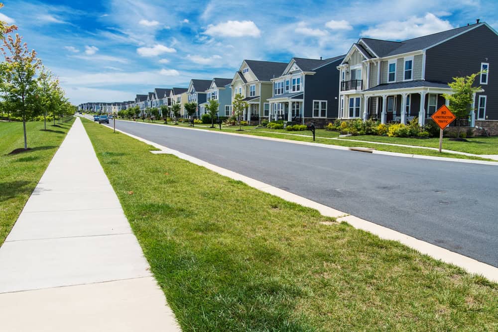 Suburban street view with a row of modern houses maintained by HOAs, a grassy lawn, and a sidewalk under a clear blue sky. A caution sign is visible on the right.
