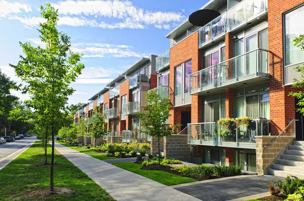 A row of modern red brick apartments with glass balconies, meticulously maintained by attentive property managers, features neatly landscaped surroundings, trees, and a sidewalk basking in the sunlight.
